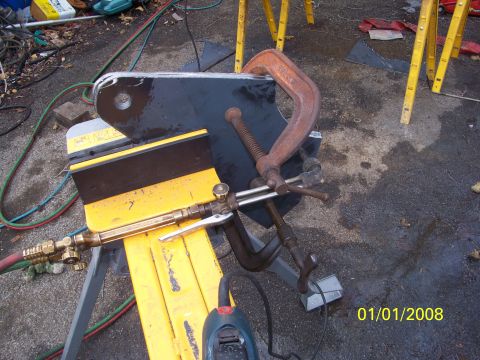 1989 Ford Bronco - Fabrication in progress: a black steel bracket plate with a large mounting hole clamped by two C-clamps on a yellow jack stand and work sup…