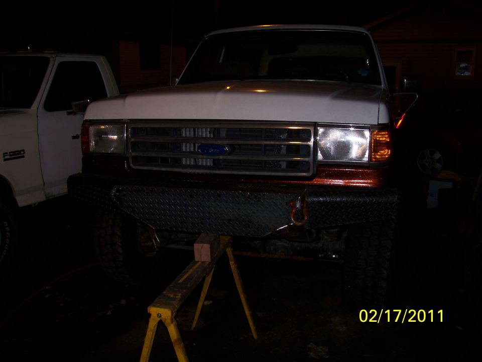 Front view of a white Ford F-250 truck with a custom bumper, supported by jack stands. Another white Ford truck is partially visible to the left