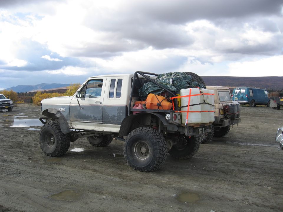 1990 Ford F-250 pickup truck modified with large tires and bed rack carrying cargo, parked with other vehicles