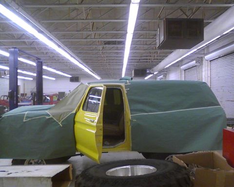 1978 Ford Bronco - A yellow truck is partially covered in a garage, with a spare tire and box in the foreground.