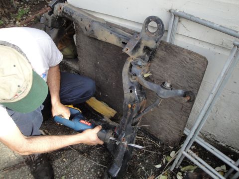 1995 Ford Taurus - A person uses a reciprocating saw to cut a rusty car subframe assembly outdoors.