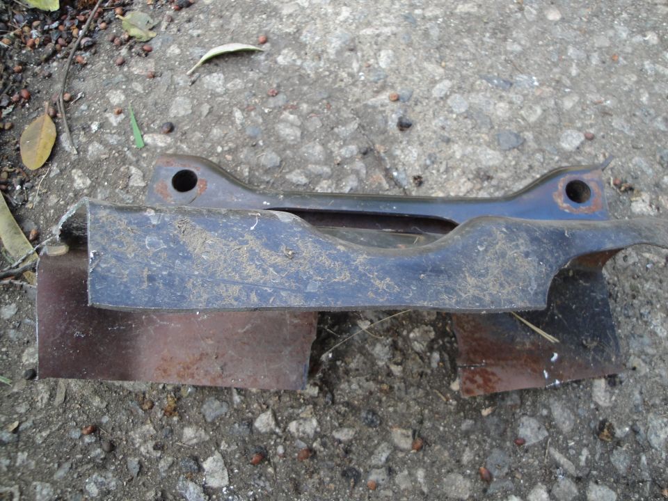 Metal subframe component with two mounting holes, resting on rusty metal pieces