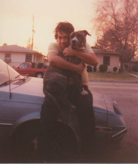 1968 Ford Mustang - A man embraces his dog while sitting on the hood of a car in a suburban setting.
