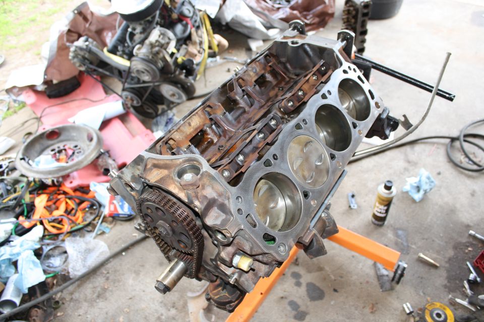Engine block with pistons visible, on an engine stand. Other engine parts and tools are scattered around
