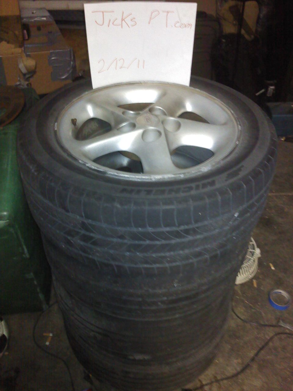 Stack of four tires with a silver five-spoke wheel on top, a handwritten note behind it