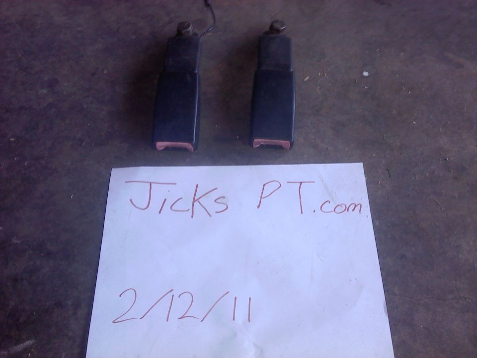Two black seatbelt buckles with pink tongues on a dark surface, with a paper showing "Jicks PT.com" and "2/12/11"