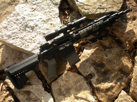 1996 Ford Bronco - An assault rifle with a scope and tactical grip is placed on a rocky surface.