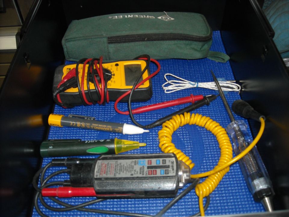 Open tool case with various electrical testing tools including multimeters and voltage testers on a blue mat