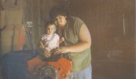 1979 Chevrolet Camaro - A man holds a baby while posing with an orange engine block in what appears to be a garage.