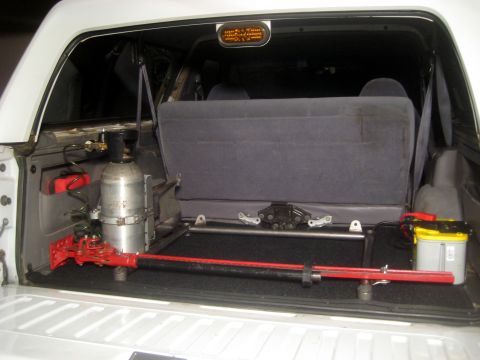 1996 Ford Bronco - The cargo area of a vehicle contains a red jack, a silver air tank, and a yellow battery, suggesting aftermarket modifications.