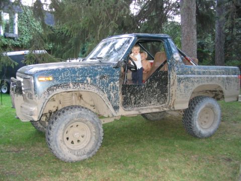 1986 Ford Bronco - Mud-covered 1986 Ford Bronco with two children in the cab, parked on grass.