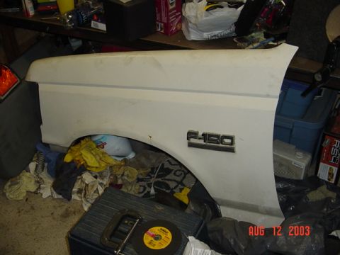 1987 Ford Bronco - A white Ford front fender with a chrome F-150 badge leans against a workbench in a cluttered garage, showing the wheel arch and body-side c…