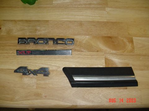 1987 Ford Bronco - Four removed Ford Bronco exterior trim pieces laid out on a butcher-block table: a chrome BRONCO script badge, a red-lettered XLT badge wit…