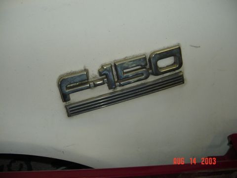 1987 Ford Bronco - Close-up of a chrome and blue F-150 fender emblem with ribbed chrome underline on a dusty white body panel, a red stripe visible along the…