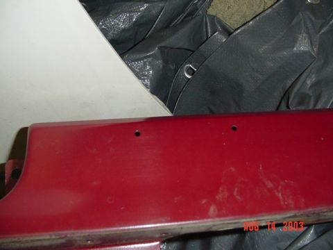 1987 Ford Bronco - Detail of a red-painted body panel edge with two small trim mounting holes and light scratches, laid over a gray tarp next to a white panel…