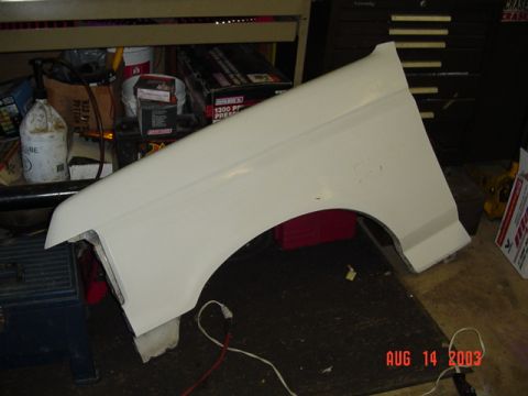 1987 Ford Bronco - A white truck front fender removed from the vehicle and propped upright on a workbench in a garage, showing the wheel arch, headlight openi…