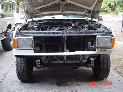1987 Ford Bronco - Head-on view of a white Ford truck in a driveway with the hood open and the grille removed, exposing the radiator, core support and wiring;…