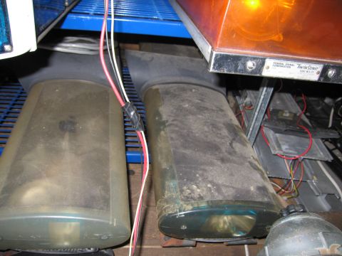 2001 Ford Crown Victoria - Close-up of dusty emergency light bars stored on a shelf, including two clear/smoked domed units and an amber-lensed Federal Signal Corpora…