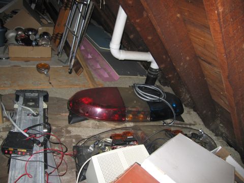 2001 Ford Crown Victoria - Two police light bars stored in a dusty attic space with exposed rafters and PVC pipe: the upper unit has red, amber and blue lens sections…