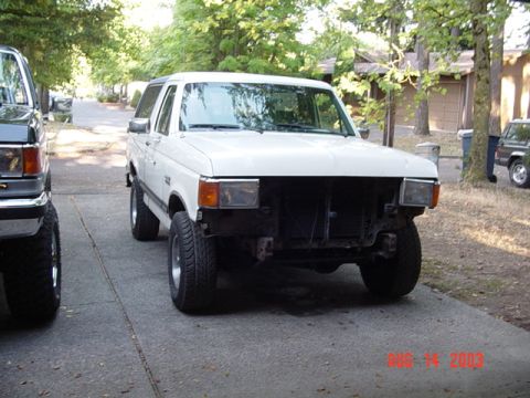 1987 Ford Bronco - Three-quarter front view of a white late-'80s Ford Bronco with removable hardtop, parked on a tree-shaded driveway with its grille, bumper…