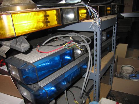 2001 Ford Crown Victoria - Stacked emergency light bars on grey steel shelving: an amber-and-clear lens bar on the upper shelf and two blue-lensed bars with clear tak…