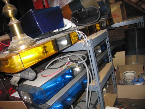 2001 Ford Crown Victoria - A collection of emergency light bars are stored on metal shelving units, with various boxes and other items in the background.