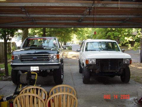 1987 Ford Bronco - Two lifted late-1980s Ford trucks parked nose-in on a driveway, shot from inside a garage: a black one at left with chrome bumper and Orego…