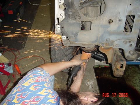 1987 Ford Bronco - Overhead shot of a person in a blue flame-print shirt using an angle grinder on the stripped front core support of a truck, throwing a brig…
