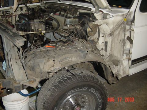 1987 Ford Bronco - Close view into the passenger-side engine bay of a white truck with the fender removed, exposing the dusty inner fender apron, firewall, br…