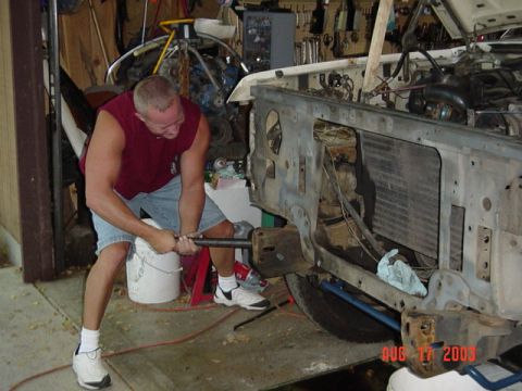 1987 Ford Bronco - A man in a maroon sleeveless shirt and denim shorts leans into a long pry bar at the stripped front end of a white truck, working against t…