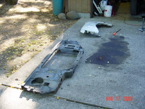 1987 Ford Bronco - A truck's front radiator core support and inner-fender assembly lying face-up on a driveway after removal, showing the headlight buckets, b…