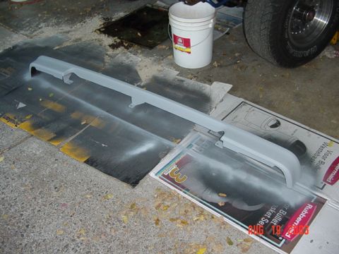 1987 Ford Bronco - A long body panel, likely a valance or grille surround, freshly coated in gray primer and laid on cardboard and a 3M sanding-sheet box on a…
