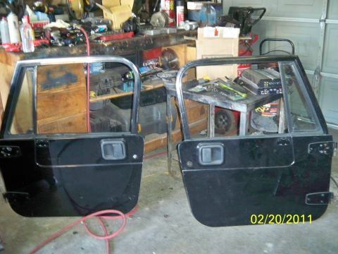 1992 Jeep Wrangler - Two black Jeep Wrangler doors are displayed in a workshop setting, with tools and equipment in the background.