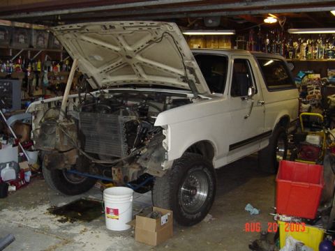 1987 Ford Bronco - Full side-front view of a white Ford Bronco up on jack stands in a cluttered garage with the hood open and the front clip, grille, bumper,…