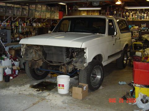 1987 Ford Bronco - Driver-side three-quarter view of a white full-size Ford Bronco in a workshop with its front clip disassembled, grille, headlights and fron…