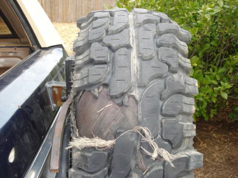 1985 Ford Bronco - A severely damaged tire, revealing its internal construction, is mounted on the rear of a dark-colored vehicle.