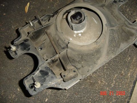 1987 Ford Bronco - Back side of a dusty removed sealed-beam headlight housing/bucket lying on a concrete floor, showing the molded plastic mounting flanges, w…