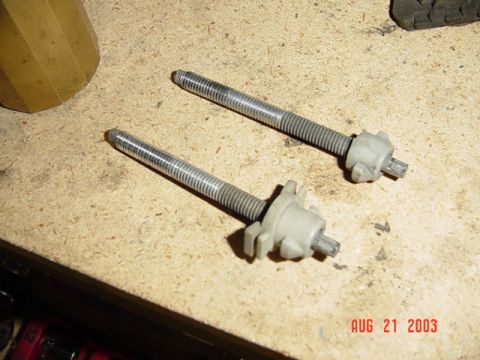 1987 Ford Bronco - Two headlight aim-adjuster screws with long threaded shafts and white nylon gear housings lying on a particleboard workbench. The hardware…