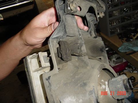 1987 Ford Bronco - A hand holds a dusty removed headlight assembly with its metal mounting bracket and adjuster screws visible from the back side, over a work…