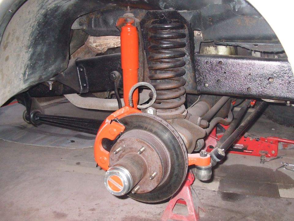 Close-up of vehicle's front suspension, brake rotor, caliper, and hub assembly. Orange shock absorber and brake caliper visible