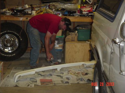 1987 Ford Bronco - A man in a red shirt, jeans and ball cap bends over a removed body panel laid on newspaper in a garage, working with a spray can beside a w…