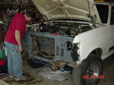 1987 Ford Bronco - A man in a red shirt and jeans works on the stripped front end of a white Ford Bronco in a garage, with the hood raised and the front fende…