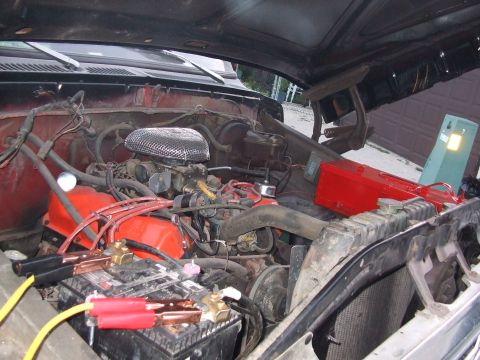 1979 Ford Bronco - The engine bay of a red Ford Bronco shows jumper cables connected to the battery under an open hood.