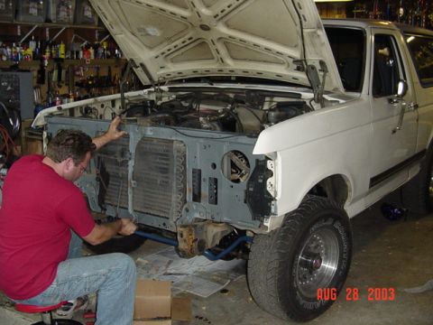 1987 Ford Bronco - A man in a red shirt sits on a low shop stool working at the exposed core support of a white Ford Bronco with the hood up and the grille, b…
