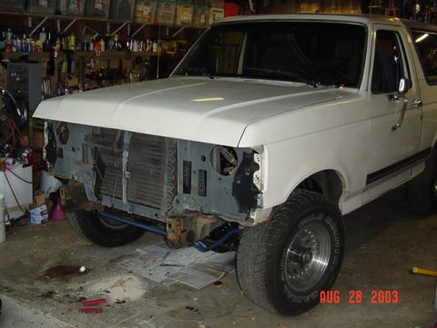 1987 Ford Bronco - Three-quarter front view of a white Ford Bronco in a garage with the hood closed but the grille, bumper and headlight assemblies removed, l…