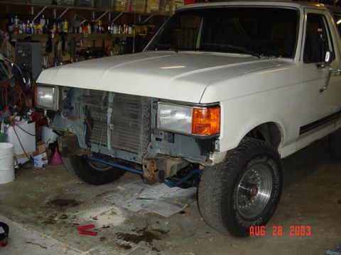 1987 Ford Bronco - Three-quarter front view of a white late-'80s Ford truck in a home garage with the grille and bumper off, showing a freshly primered gray c…