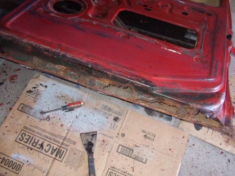 1979 Ford Bronco - A heavily rusted car door sits on cardboard with a screwdriver and scraper.