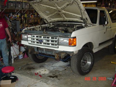 1987 Ford Bronco - White late-'80s Ford truck in a cluttered home garage with the hood propped open and the refinished white grille with blue FORD lettering j…