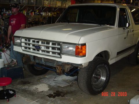 1987 Ford Bronco - White 1980s Ford Bronco sitting in a home garage with the front bumper removed, exposing blue-painted frame horns and the bare front crossm…