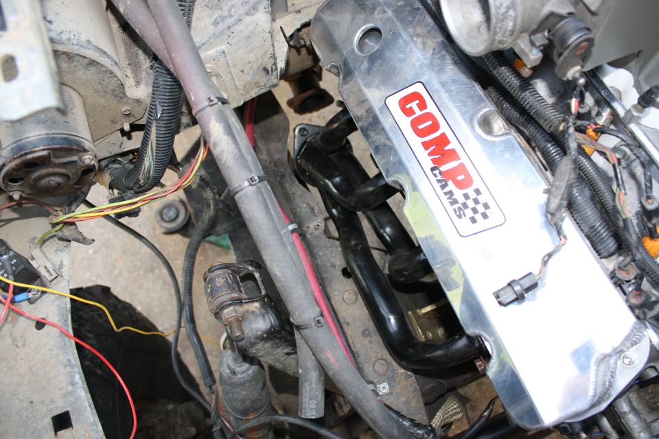 Polished Comp Cams valve cover and black tube headers in a dusty engine bay